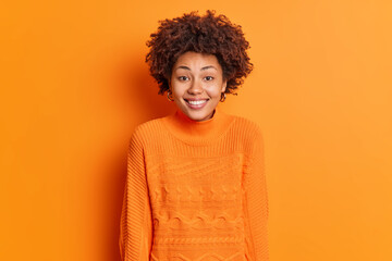 Portrait of happy young African American woman smiles broadly has white perfect teeth expresses joy wears casual sweater isolated over orange background. People emotions and feelings concept.