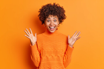 Happy surprised curly haired African American woman raises palms looks with amazement at camera...