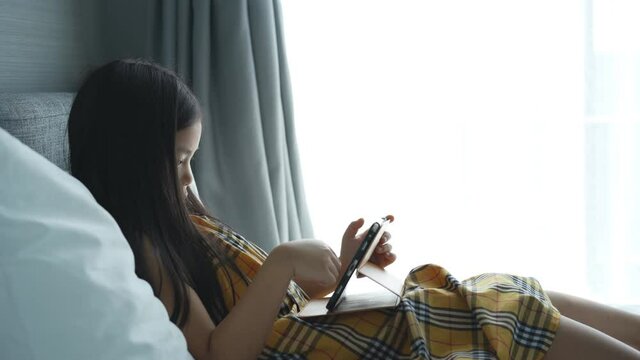 Asian child or kid girl learn from home or study online and e-learning on computer tablet to communication and leaning against the pillow on bed or people play game in bedroom and white window curtain