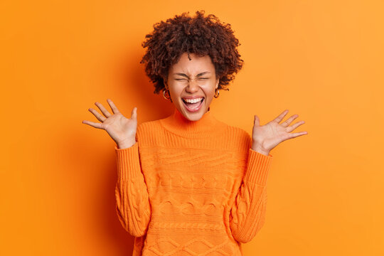 Positive overjoyed young African American woman raises palms feels very glad expresses joy dressed in casual jumper isolated over orange background. People emotions happiness concept. Monochrome