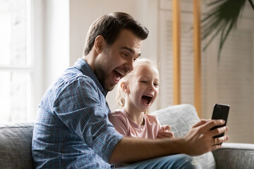 Excited dad and daughter girl sitting on couch, using mobile phone for video call, shouting and laughing at screen. Happy father and kid having fun together at home, taking selfie on smartphone