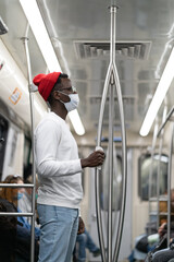 Fototapeta na wymiar African American hipster man in glasses and red hat wear face mask in subway train during covid-19 pandemic, holding handrail through the napkin. Coronavirus, new normal concept. Defocused.