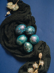 Mother of pearl Easter eggs lie on a dark fabric and a blue background. The fabric flows in a wave....