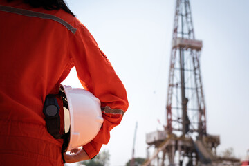 A worker is holding safety hardhat or helmet with blurred background of drilling rig derrick...