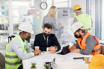 Professional designers, engineers and architects wearing medical masks and helmets analysing blueprints of new building. Diverse colleagues sitting at office and working on common project.