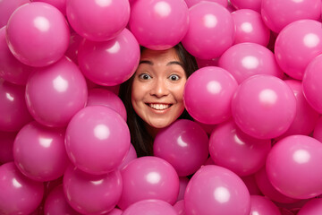 Fototapeta na wymiar Positive brunette Asian woman smiles gently being in good mood poses through heap of inflated pink balloons prepares for birthday celebration shows white teeth. People holiday partying concept