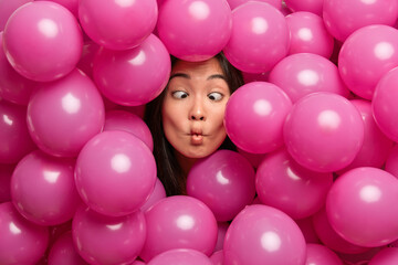 Fototapeta na wymiar Funny childish young Asian woman makes fish lips crosses eyes foolishes around while decorating hall with inflated balloons for special occasion has crazy weird face. People and holidays concept
