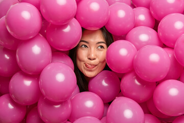 Fototapeta na wymiar Photo of dreamy Asian woman licks lips with tongue concentrated above thoughtfully surrounded with inflated pink balloons thinks how to celebrate special occassion in life. Festive event concept