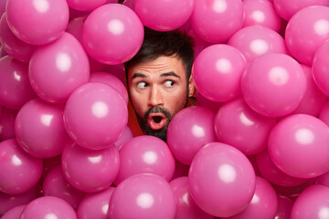 Obraz premium Surprised bearded European man sticks head through decorated background with inflated balloons prepares for holiday being shocked by unexpected relevation. People celebration emotions concept