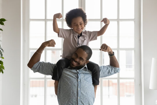 Happy funny kid riding on dads shoulders. African American daddy wearing glasses and son playing together, showing hand strength, flexing biceps, laughing and having fun. Family home activity concept