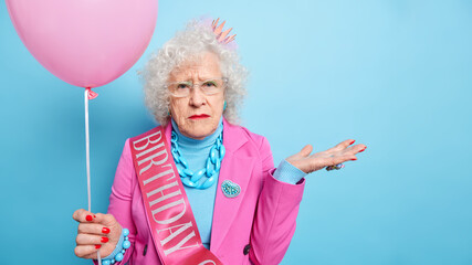 Studio shot of wrinkled grey haired elderly woman raises palms looks seriously at camera dressed in festive clothes holds inflated balloon celebrates anniversary isolated over blue background