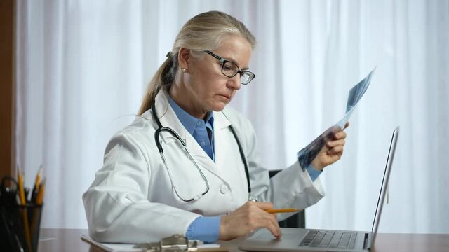 Female doctor wearing lab coat and stethoscope looks at X-ray image of patient. Concept of woman physician working in healthcare in office.