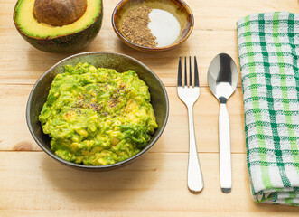 Fresh guacamole on a dish placed on a wooden table with ingredients for homemade guacamole: avocados, lemon, salt, and pepper. Top view.  Concept of traditional Mexican preparation