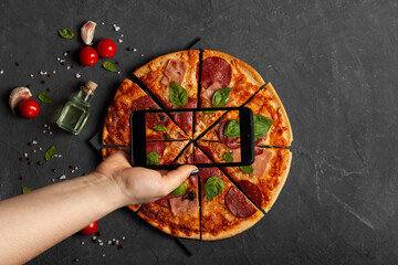 Young woman with a phone takes a picture of a fresh pepperoni pizza on a concrete table with cooking ingredients cherry tomatoes, basil and olive oil. Top view.