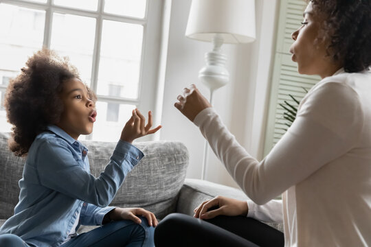 Excited African American kid talking to mom nonverbal. Mother training girl to communicate with hand and finger gestures. Teacher using sign language for giving lesson to deaf disabled child at home