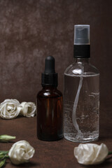 Glass bottles with facial toner and oil serum placed on table with rose flowers on brown background in studio 