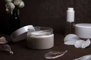Hydrating facial cream in plastic jar placed with flower petals on table on dark background in studio for concept of beauty treatment and skin care routine 