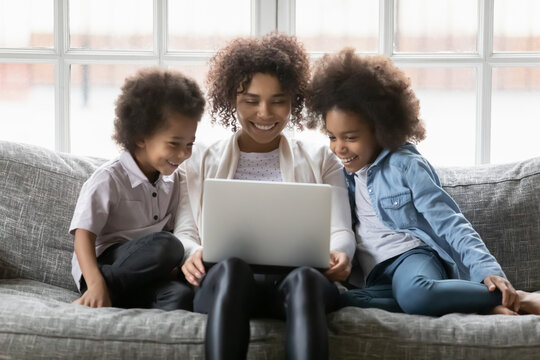 Happy mom and two children using laptop for video call, shopping on internet, watching funny movie online. Cute African American family resting on couch at home, looking at computer screen together