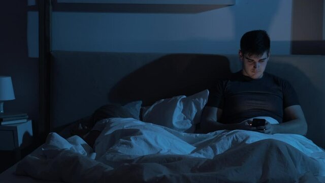 Online cheating. Marriage problem. Secret affair. Disloyal husband using dating app on mobile phone in bed with sleeping wife late at night in dark blue light.