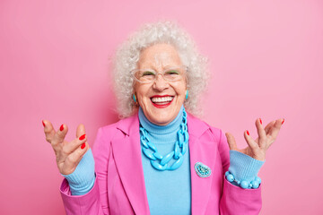 Happy senior curly woman enjoys life in old age raises hands smiles positively dressed in...