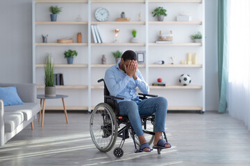 Depression and loneliness in disabled adults. Young black guy in wheelchair crying at living room,...