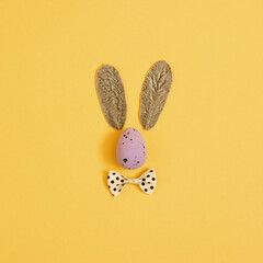 Easter egg with bow tie and easter bunny ears made of leaves. Layout on yelow background