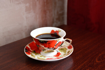 Elegant coffee service with red poppies. A cup of black coffee on the wooden table against the background of red curtain and beige wallpaper.
