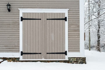 Two grey wooden exterior barn style doors with black wrought iron hinges in an old vintage  building. The building has been painted grey with white trim. The foundation has a rock foundation.