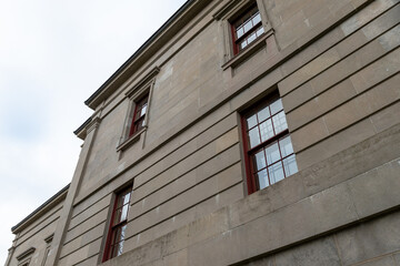 The exterior of a vintage limestone block wall with multiple windows. The tall government building has a blue sky with clouds in the background. The trim around the double hung windows is red wood