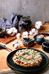 Homemade japanese fast food okonomiyaki cabbage pancake with onion, pickled ginger, mayo sauce on black ceramic plate. Chopsticks, teapot, cotton, ingredients above. Texture background. Flat lay