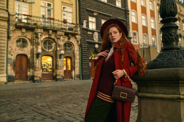 Elegant fashionable redhead woman wearing dark red fedora hat, stylish midi coat, knitted vest, holding small leather bag, posing in street. Copy, empty space for text