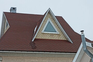 brown brick attic with one triangular window on the tiled roof of a private house against a gray sky