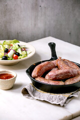 Grilled italian sausages salsiccia in cast-iron pan served with tomato sauce and plate of fresh vegetable salad on white marble table. Balanced dinner