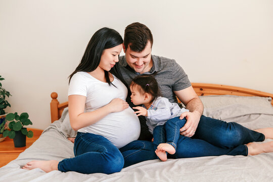  Family Asian Chinese pregnant woman and Caucasian man with toddler girl sitting on bed at home. Mother, father and baby daughter expecting waiting for a new family member. Ethnic diversity.
