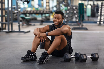 Fototapeta na wymiar Smiling black muscular man having break while exercising at gym