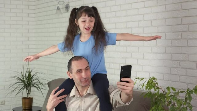Fly with dad. The daughter sits on the shoulders of her father and flies, and the dad takes pictures with his smartphone.