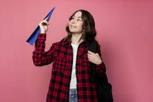 Smiling young woman in red check shirt with black backpack holding paper airplane on pink background. Girl tourist is throwing origami plane. Travel, journey, adventure concept. Studio shot