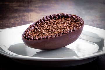 Stuffed chocolate easter egg with grated chocolate on the top and around on a white plate on a wood table.