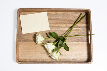 A bouquet of three white roses ion a wooden tray with an evelope.