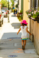 A pretty little girl wearing short summer dress, sandals and pink wide brimmed solar hat is walking alone in the shade at a sunny hot summer day. Selective focus image shows her from behind.