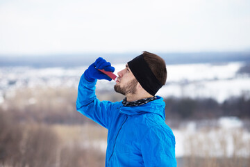 Young handsome sportsman man with a beard goes in for sports on the nature in winter. He drinks isotonic and eats sports nutrition in a tube to recuperate.