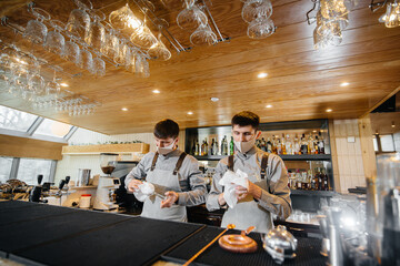 Two stylish bartenders in masks and uniforms during the pandemic, rub glasses to shine. The work of restaurants and cafes during the pandemic.