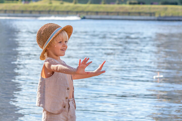 Happy little blond caucasian boy wearing straw hat stands by the river. Cheerful smiling boy lifts his hands. Copy space for your text. Summer kids fun theme.