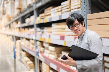 Young Asian man worker doing stocktaking of product in cardboard box on shelves in warehouse by using clipboard and pen. Physical inventory count concept