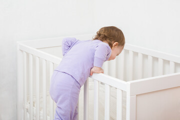 Baby girl in purple pajama trying to get in white crib and going to sleep. Toddler development.
