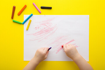 Baby hands holding wax crayons and drawing first scratches lines on white paper on bright yellow table background. Closeup. Toddler development. Point of view shot. Top down view.