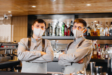 Two stylish bartenders in masks and uniforms during the pandemic, stand behind the bar. The work of restaurants and cafes during the pandemic.