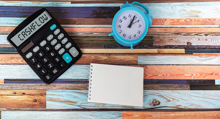 Calculator with the words Cash Flow, an alarm clock stands next to them, a notepad for writing on a wooden table