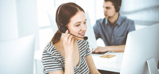 Casual dressed young woman using headset and computer while talking with customers online. Group of operators at work. Call center, business concept