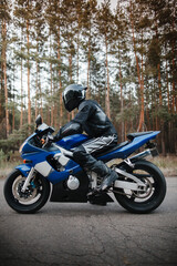 Fototapeta na wymiar Motorcyclist in leather protective suit and black helmet sits on sports motorcycle. Biker in black rides on the road against the background of the forest. Side view